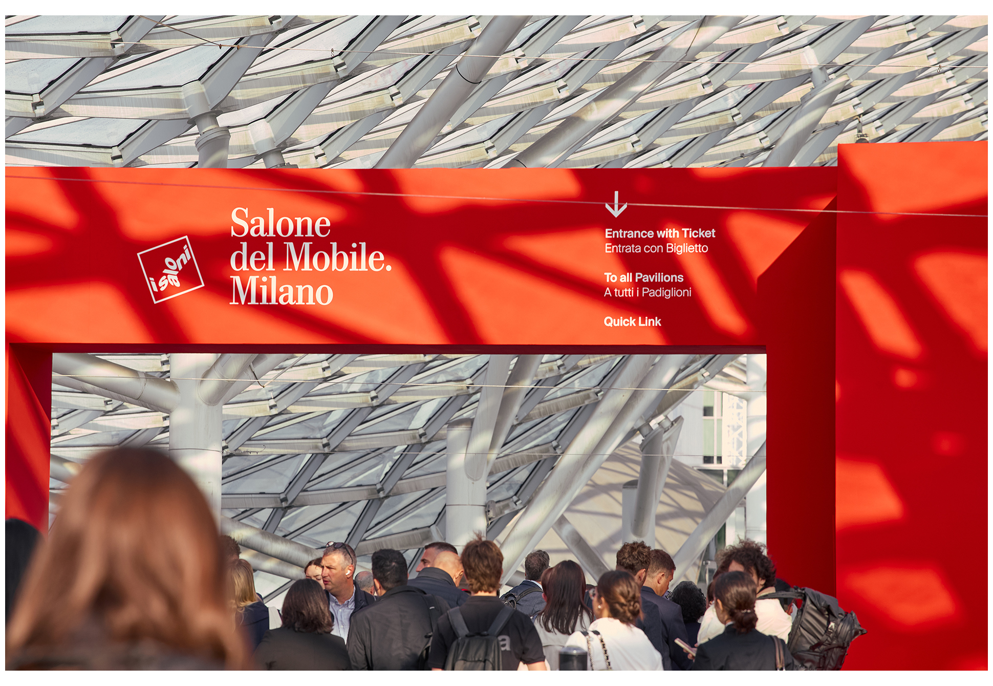 Visitors passing in front of a red structure installed at the entrance of the Milan Salone del Mobile venue