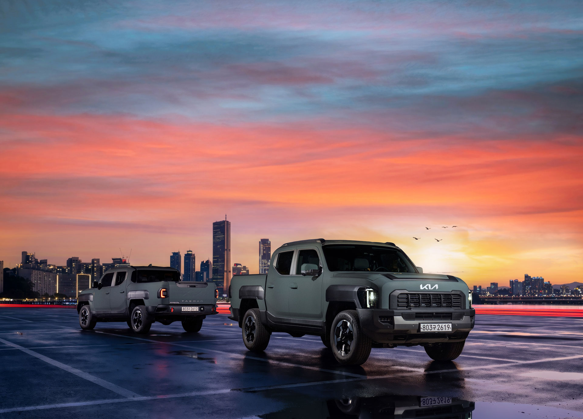 Two Kia Tasman pickup trucks on wet pavement against a sunset city backdrop