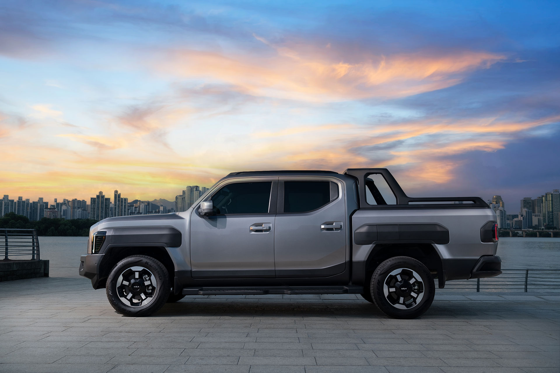 Side view of a silver Kia Tasman pickup parked in a riverside plaza bathed in sunset light