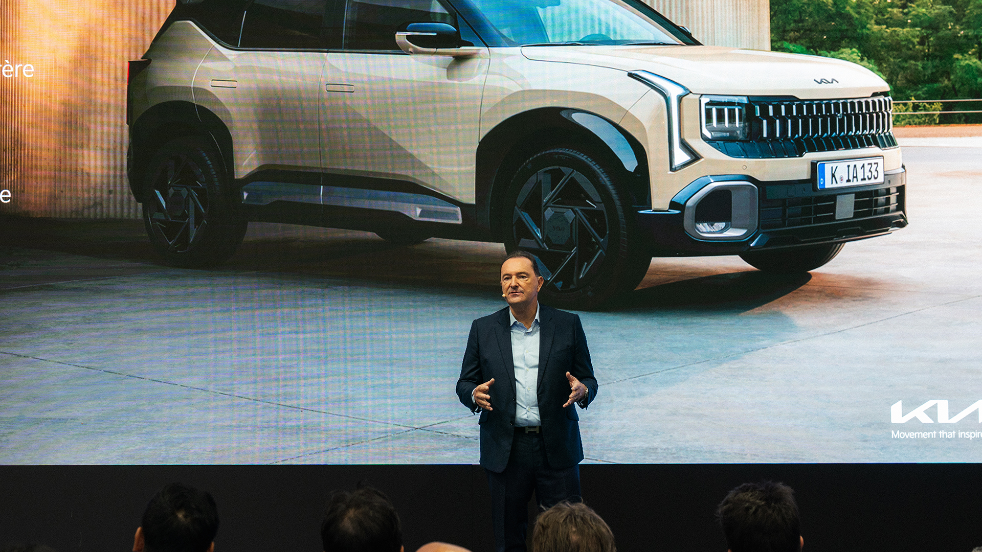 Presenter before beige Kia SUV display with audience silhouettes