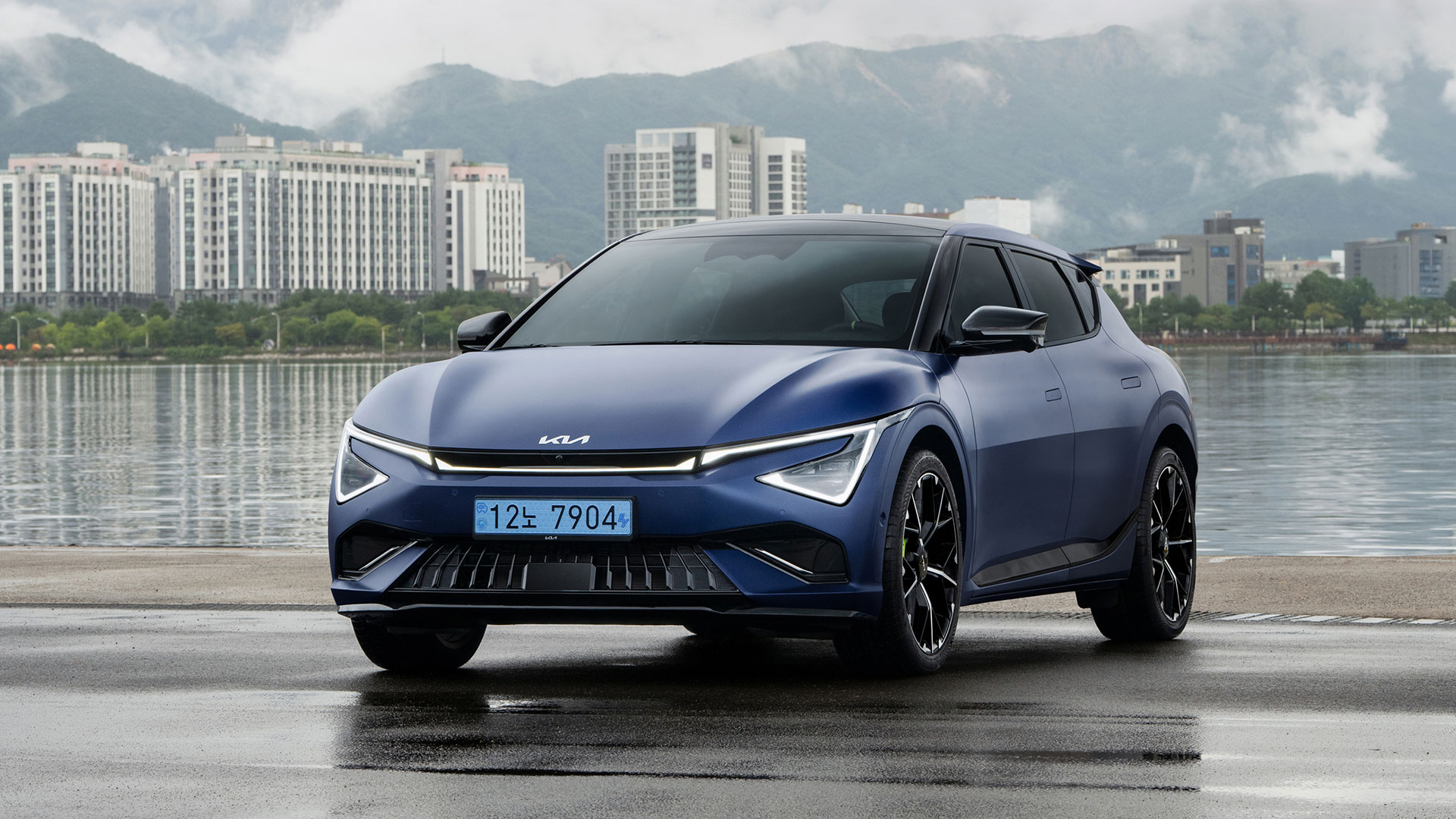 Front view of a matte blue EV6 GT parked on wet pavement against a backdrop of misty mountains and a city